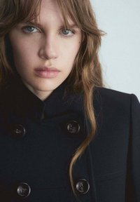 Navy double-breasted coat featuring large black buttons, structured collar, and smooth fabric, complemented by long, wavy hair.