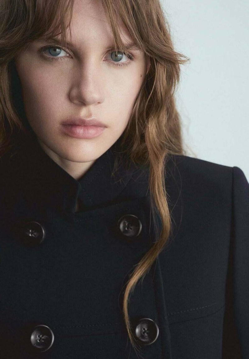 Navy double-breasted coat featuring large black buttons, structured collar, and smooth fabric, complemented by long, wavy hair.