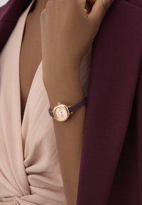 Zeltīta apaļa pulksteņa ar rozā kloņu un sudraba rādītājiem, kuram ir purpursarkana silikona siksna. Valkāts uz rokas pret pelēku žaketi.