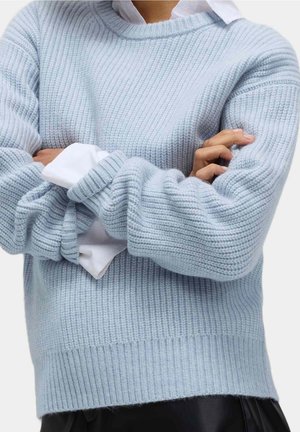 Personne portant un pull en maille bleu clair par-dessus une chemise blanche, bras croisés et pantalon noir, visible du milieu du torse jusqu'au cou.