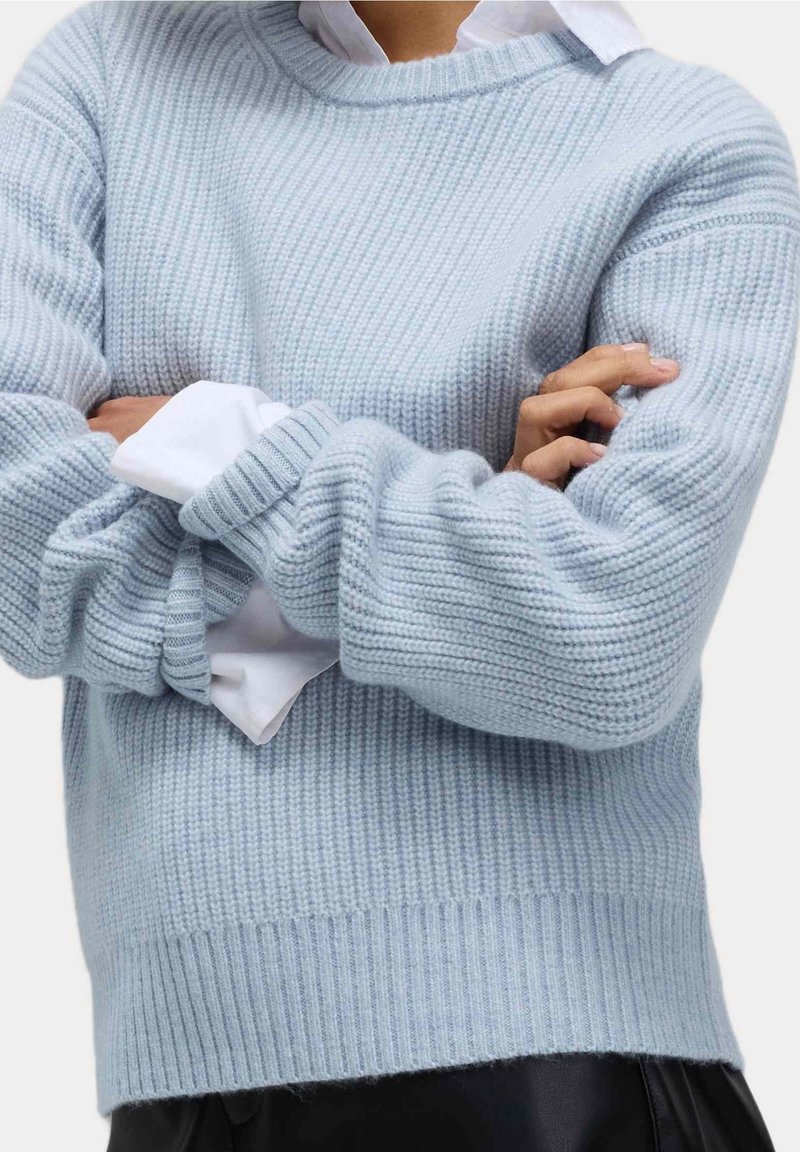 Personne portant un pull en maille bleu clair par-dessus une chemise blanche, bras croisés et pantalon noir, visible du milieu du torse jusqu'au cou.