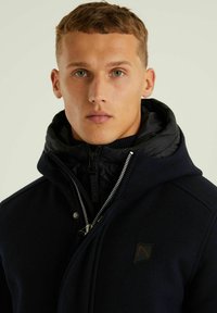 Tamsiai mėlynas vilnos mišinio paltas su juodu pūkuotu kapišonu, priekyje užtrauktukas ir sidabriniai dirbiniai; turi logotipo detalę kairėje krūtinės pusėje.