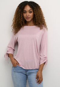 Ljusrosa blus med trekvartsärmar, rynkade knytdetaljer vid handlederna, mjuk textur och avslappnad passform, kombinerad med ljusblå jeans.