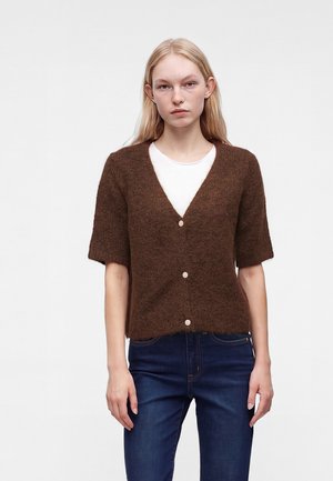 Jeune femme aux longs cheveux blonds porte un cardigan marron à manches courtes boutonné par-dessus une chemise blanche et un jean bleu foncé sur un fond uni.