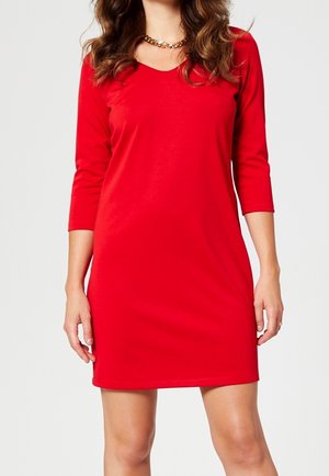 Frau in einem roten Kleid mit dreiviertel Ärmeln und einer Länge über dem Knie sowie einer massiven goldenen Halskette, die vor einem schlichten hellen Hintergrund steht.