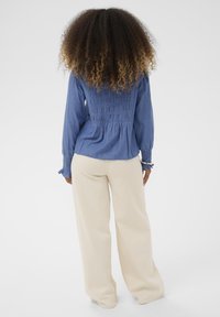 Sininen denimpaitapusero, jossa on kokoonkiristys takana ja pitkät hihat, yhdistettynä leveälahkeisiin kermaisiin housuihin. Tasainen kangas koko matkalla.