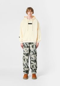 Sudadera amarilla clara con un parche negro, cordones beige, combinada con pantalones anchos estampados en verde y blanco, y zapatos marrones.