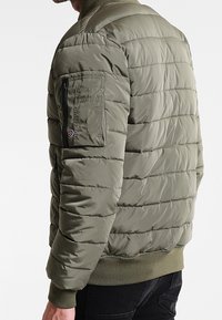 Chaqueta acolchada verde oliva con costuras horizontales, puños acanalados y un bolsillo de manga con cremallera. Textura suave y acolchada.