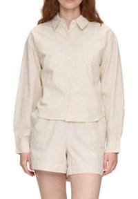 Mujer vestida con una camisa de botón de color crema, con finas rayas, y pantalones cortos a juego, de pie con las manos relajadas a los lados.