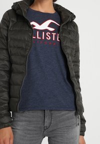 Ciemnozielona kurtka puchowa z zamkiem błyskawicznym i kapturem, noszona na granatowej koszuli z białym i czerwonym tekstem graficznym, dopełniona szarymi dżinsami.