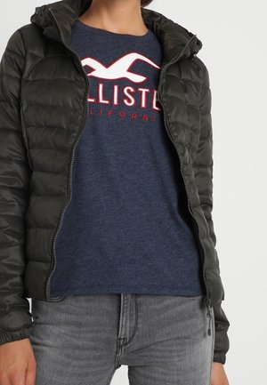 Mørkegrønne dunjakke med lynlås og hætte, båret over en marineblå skjorte med hvid og rød grafisk tekst, suppleret med grå jeans.