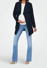 Zwangerschapsjas in donker marineblauwe stof met een open voorkant, gelaagd ontwerp. Gecombineerd met lichtblauwe uitlopende jeans en beige spitse hakken.