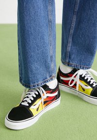 Baskets noires avec des motifs de flammes rouges et jaunes. Lacets blancs et semelles en caoutchouc durables. Portées avec un jean en denim bleu sur une surface verte.