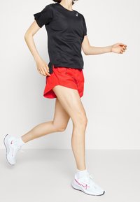 Camiseta deportiva negra, pantalones cortos rojos con aberturas laterales y zapatillas blancas con acentos rosas. Tejido texturizado, corte ceñido, diseño de alto rendimiento.