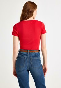Rode katoenen T-shirt met korte mouwen en ronde hals; gecombineerd met hoge blauwe jeans met een bruine riem en vier achterzakken.