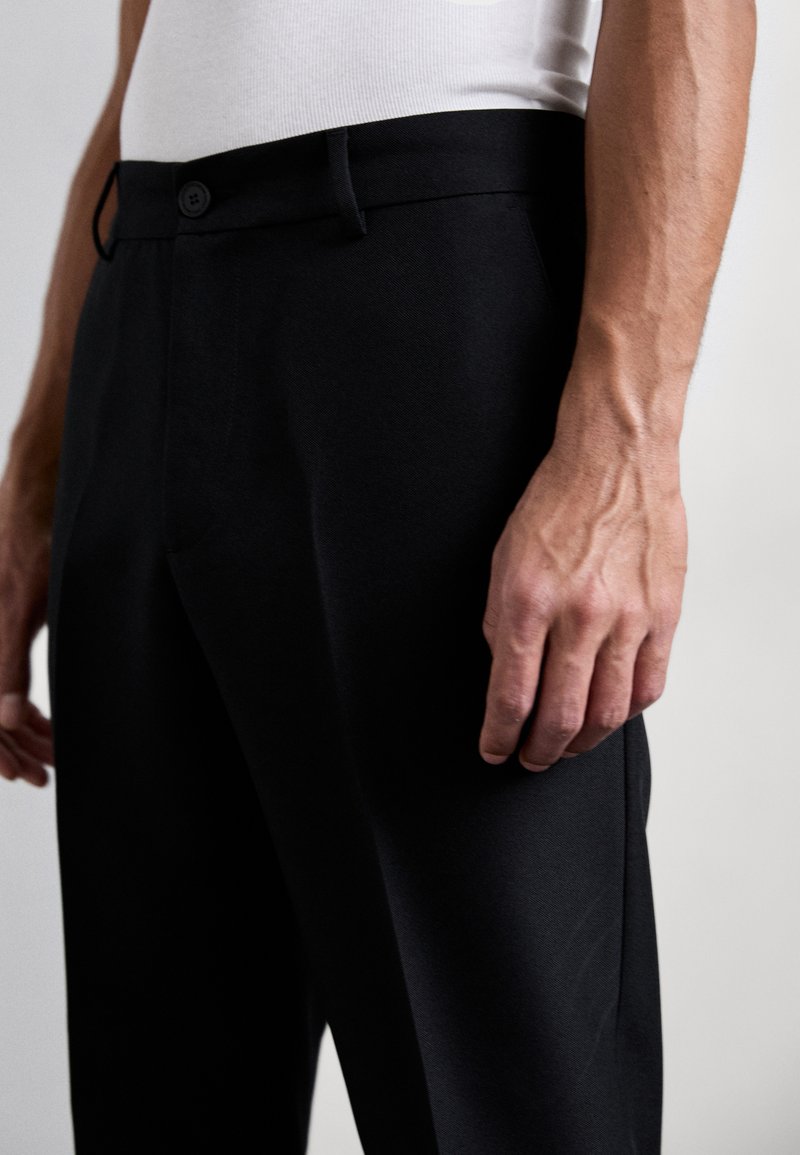 Man in zwarte op maat gemaakte broek en een witte mouwloze shirt, met zichtbare aderen op hand en arm.