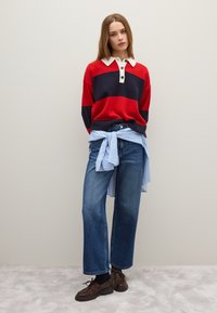 Rood en marineblauw gestreepte sweater met een crèmekleurige kraag en knopen, gelaagd over een blauw gestreepte blouse, gecombineerd met een wijde denim jeans en bruine schoenen.