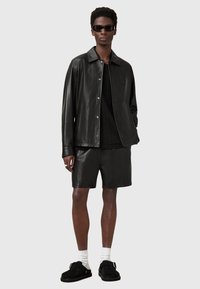 Schwarze Lederjacke mit Knopfleiste, kombiniert mit passenden Shorts. Schwarze Sonnenbrille, weiße Socken und schwarze Slipper sind ebenfalls enthalten.