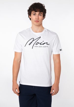 Junger Mann mit lockigem Haar, der ein weißes T-Shirt mit der Aufschrift „Moin von der Küste“ und dunkle Hose trägt, steht vor einem schlichten Hintergrund.