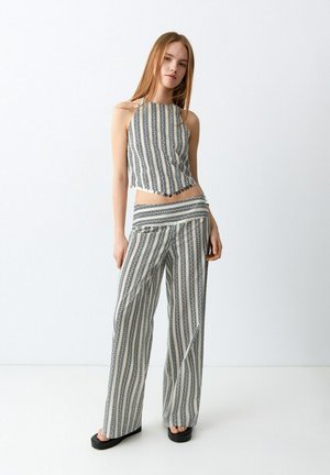 Mladá žena nosí čierno-biely pruhovaný halter top a široké nohavice, stojac na jednoduchom bielom pozadí.