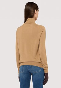 Beige polokrage tröja i mjukt stickat material, med långa ärmar och en åtsittande fåll. Kombinerad med blå denimjeans och bruna handskar.