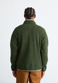 Green fleece pullover with a high collar, featuring a textured fabric and a white hem at the bottom. Worn with brown pants.