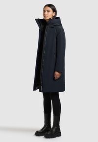 Lang marineblauwe jas met hoge kraag en ritssluiting, voorzien van zijzakken en een gladde, waterafstotende stof. Zwarte enkellaarsjes.