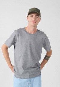 Grijs katoenen T-shirt met korte mouwen en een ronde hals, gecombineerd met lichtblauwe denim shorts. De persoon draagt een groene pet.