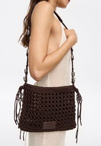 Borsa a mano all'uncinetto marrone con design testurizzato, dotata di una tracolla intrecciata decorata con hardware in argento e nappine sui lati.