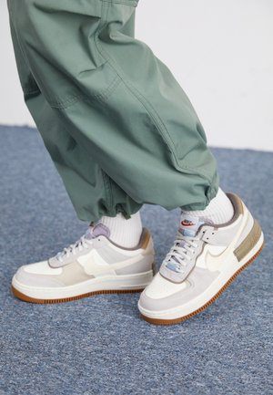 Persona con zapatillas Nike gris claro y beige con suelas de goma, calcetines blancos y pantalones verdes sueltos, de pie sobre una alfombra azul.