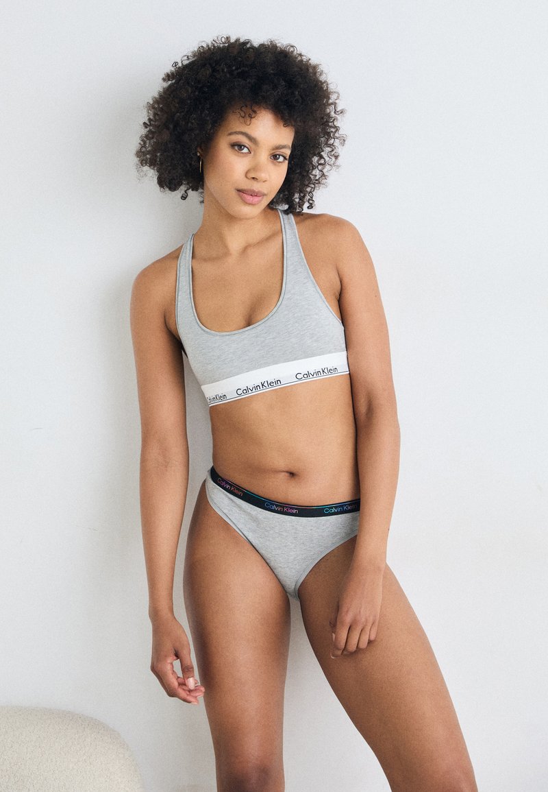 Grå bomull sport-bh med en hvit Calvin Klein-stripe og matchende grå stringtruse med en flerfarget logo-linning. Enkel, tettsittende design.