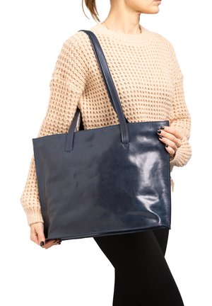 Woman in beige knitted sweater and black pants holding large navy blue leather tote bag with dual handles over shoulder.