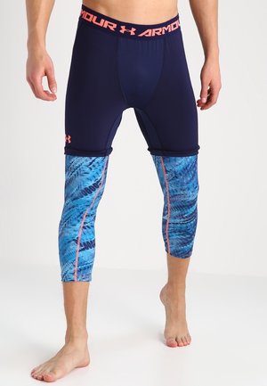 Hombre con pantalones cortos de compresión azul marino con leggings de compresión azul con patrón debajo, de pie descalzo sobre suelo blanco.