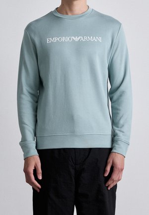 Person trägt ein hellblaues Emporio Armani Sweatshirt mit weißem Logo-Schriftzug auf der Brust und schwarze Hose vor einfarbigem Hintergrund.