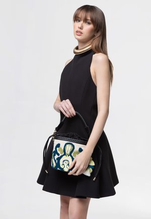 Donna con abito nero senza maniche che tiene una piccola borsa intrecciata con motivo blu, verde e crema, indossa un collare spesso in oro.