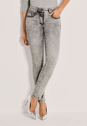Persoon draagt lichtgrijze acid-wash skinny jeans en zilveren hoge hakken, staand met één been over het andere gekruist.