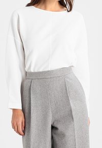 White long-sleeve top with textured fabric, paired with light grey pleated high-waist trousers made of soft wool blend.