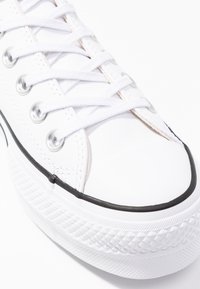 Zapatilla de lona blanca con cordones planos, ojetes plateados, puntera de goma y suela texturizada blanca con acentos negros. Vista en primer plano.
