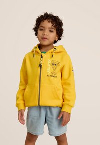Sweat à capuche jaune avec fermeture éclair, accents en matériel bleu, poches avant, et un graphique de pizza ludique ; associé à un short en denim clair.