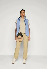 Chaleco de forro polar azul, camisa beige y pantalones caqui. Gorra beige reposando sobre un bolso negro de cinturón. Fondo liso. Ropa casual adecuada para actividades al aire libre.