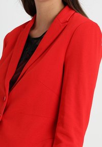 Blazer rouge avec une coupe ajustée, des revers crantés et des détails de couture subtils. Confectionné dans un tissu lisse, il présente une fermeture à un seul bouton.