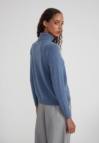 Femme aux cheveux bouclés portant un pull bleu à col roulé et un pantalon gris clair, se retournant sur son épaule devant un fond uni.