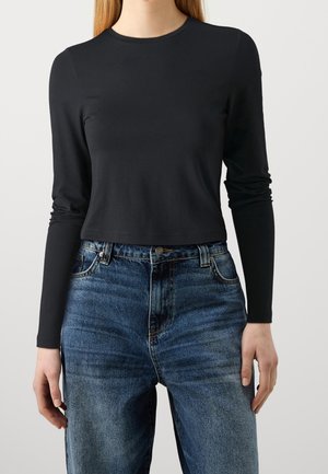 Kvinde iført en ensfarvet sort, tætsiddende langærmet top, stoppet ned i højtaljede blå denimjeans foran en hvid baggrund.