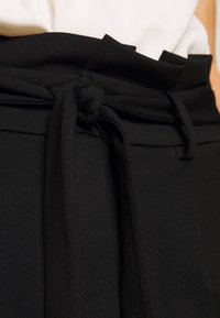 Zwarte hoogwaisted broek met een stoffen riem geknoopt, gecombineerd met een ingestopt wit topje.