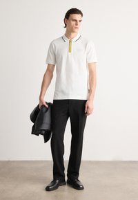 Balts polo krekls ar melnu apkakli un dzeltenu rāvējslēdzēja detaļu, apvienots ar melniem biksēm un ādas jaku, ko valkā stāvošs vīriešu modelis.
