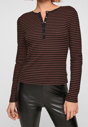 Kvinde iført en sort og brun, vandret stribet top med lange ærmer og en sort Henley-hals med knapper samt sorte bukser af kunstlæder.