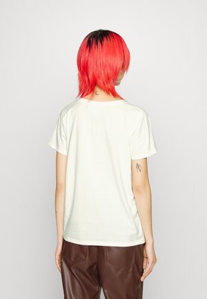 Person med kort, klart rødt hår set bagfra, iført hvid T-shirt og brune læderbukser, der viser tatoveringer på nakke og arm.