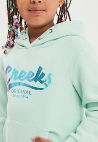 Felpa verde menta con coulisse, caratterizzata da una stampa blu lucida "Creeks Original Since 1974" sul davanti. Tessuto morbido e tasca frontale.