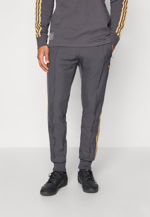Mann trägt dunkelgraue Jogginghosen mit orangenen Streifen an den Seiten und schwarzen Sportschuhen, steht vor einem einfachen hellen Hintergrund.