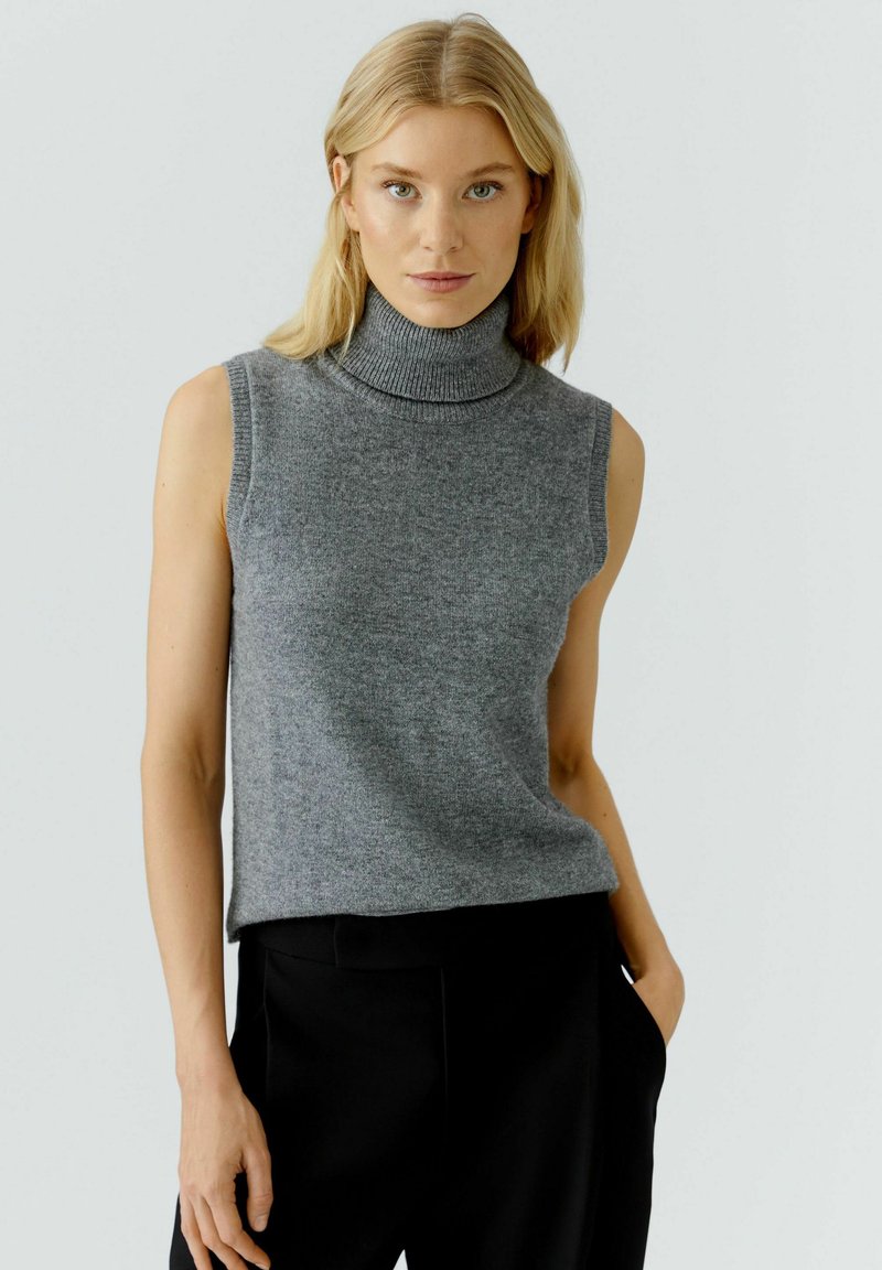 Light gray sleeveless turtleneck sweater in soft fabric, featuring a ribbed neckline and a relaxed fit. Paired with black trousers.